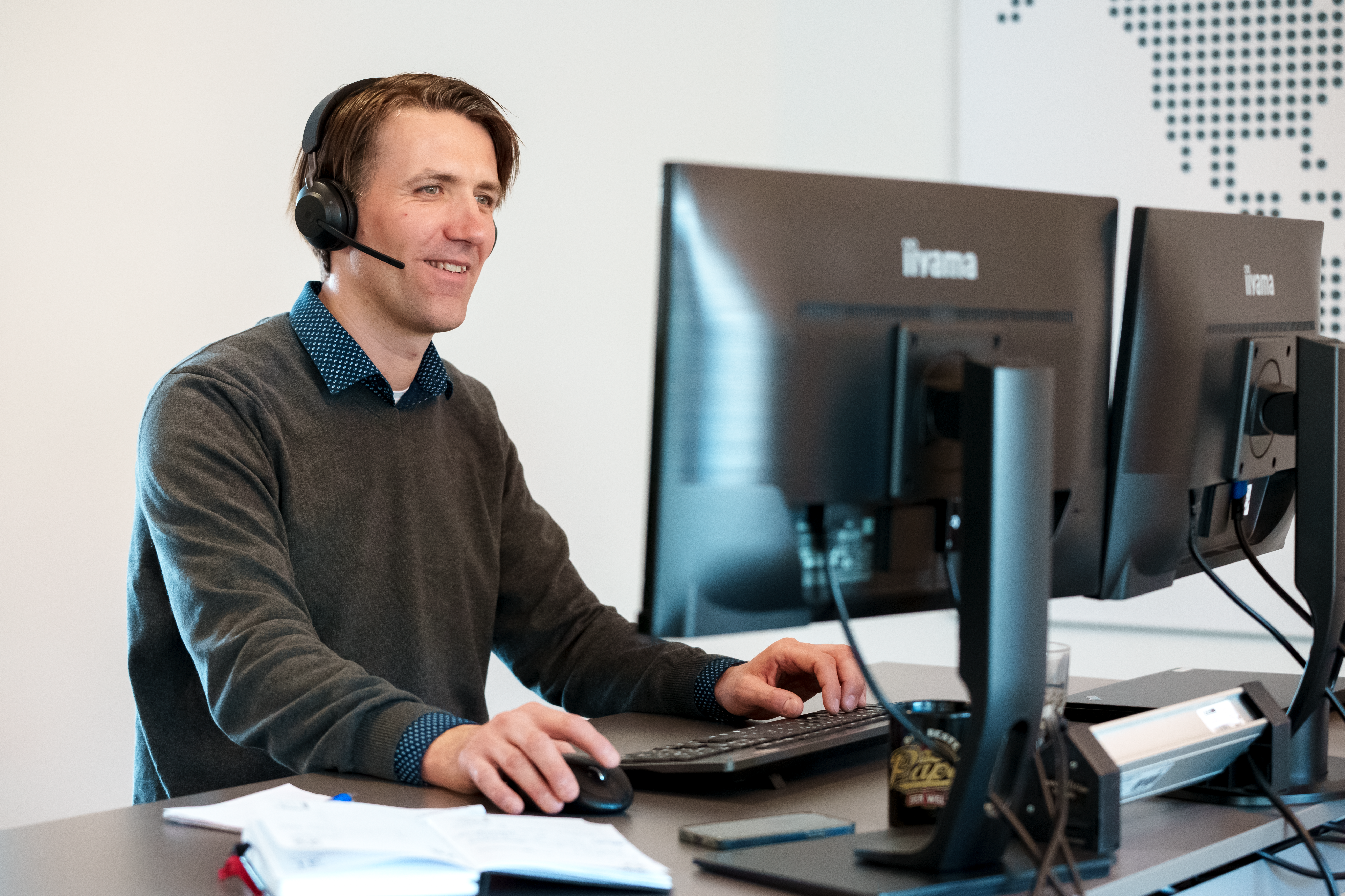 Un homme se tient debout devant son bureau, un casque sur les oreilles, et regarde son ordinateur