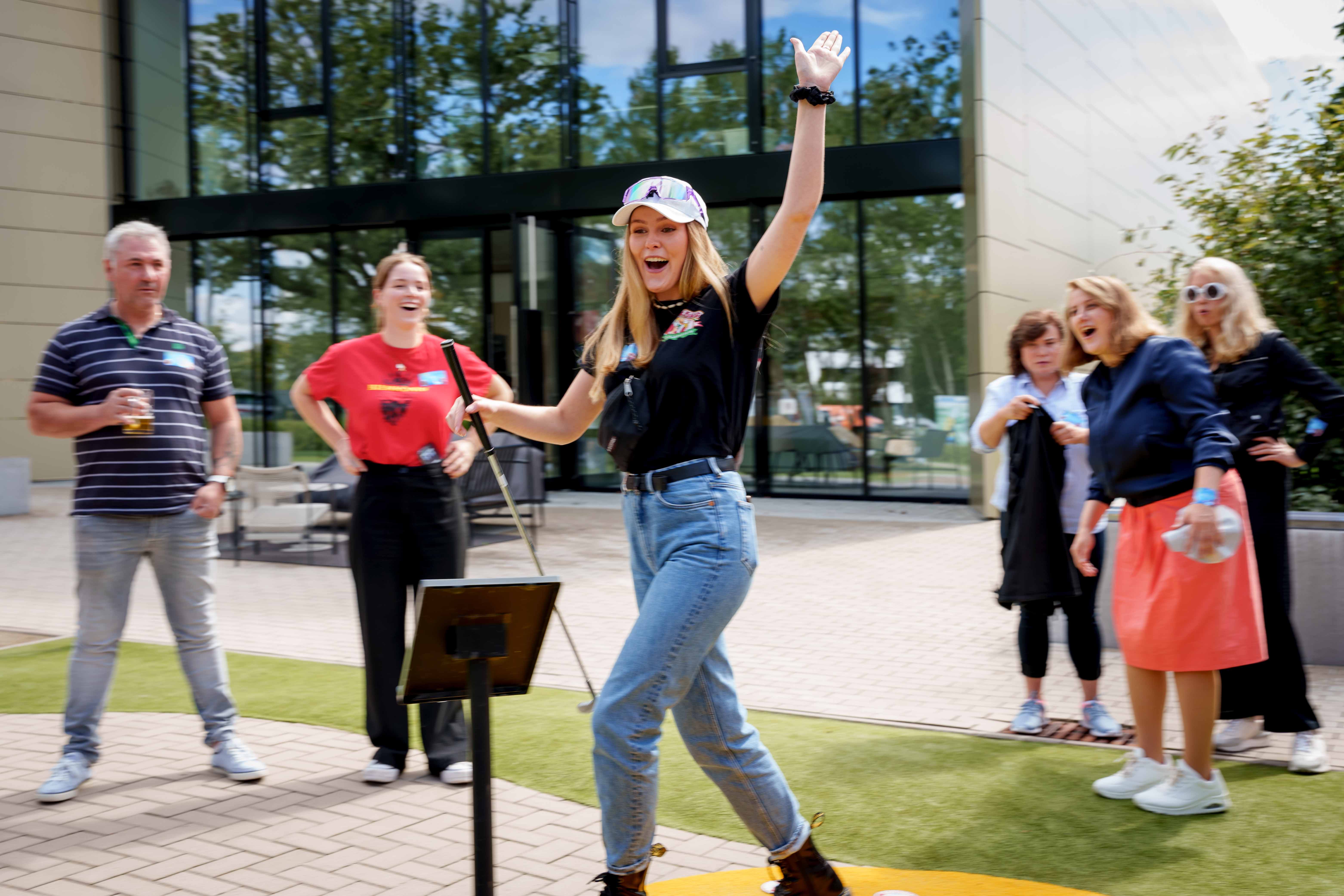 Een vrouw met een minigolfclub in haar hand juicht. Op de achtergrond zijn andere mensen blij.